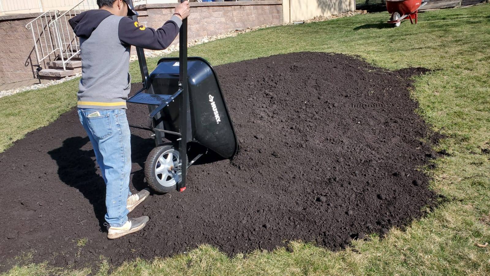 Person distributing rich soil from wheelbarrow in sunny residential yard.