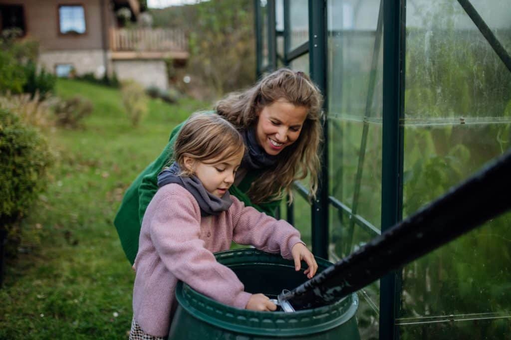 Girl and mother scooping rainwater from barell under downspout. Concept of water conservation in